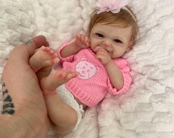 Puede incluir: Una pequeña muñeca realista de bebé con cabello castaño, que lleva un suéter rosa con un diseño de oso y una diadema blanca con una flor rosa.