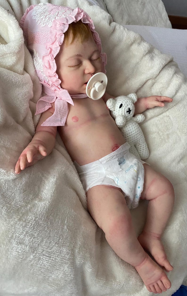 Puede incluir: Mu&ntilde;eca beb&eacute; realista con un gorro rosa con ribete de encaje y un chupete blanco. La mu&ntilde;eca, con un pa&ntilde;al, est&aacute; acostada sobre una manta blanca y sostiene un peque&ntilde;o osito de peluche blanco tejido.