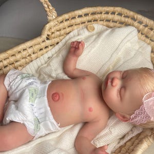 May include: A realistic newborn baby doll with light skin and blonde hair, wearing a white diaper and a pink lace headband with a bow. The doll is lying on a white knitted blanket inside a woven wicker basket with two handles and a small wooden button.