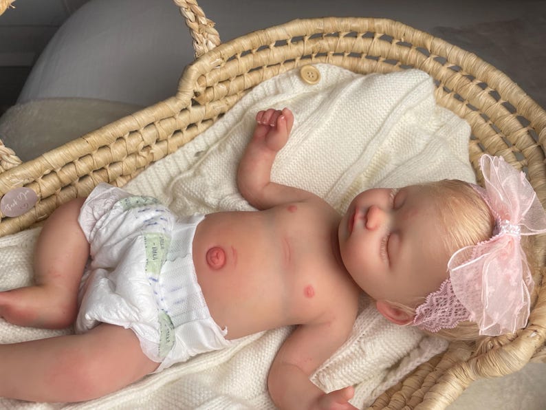 May include: A realistic newborn baby doll with light skin and blonde hair, wearing a white diaper and a pink lace headband with a bow. The doll is lying on a white knitted blanket inside a woven wicker basket with two handles and a small wooden button.