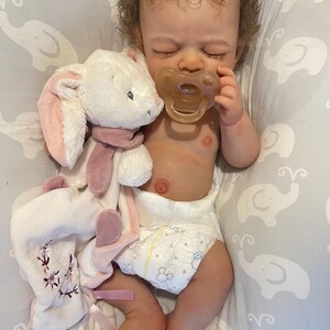 May include: A realistic baby doll with curly brown hair, wearing a nappy and holding a dummy. A white and pink plush bunny toy with a scarf is next to the doll. The doll is lying on a grey patterned surface.