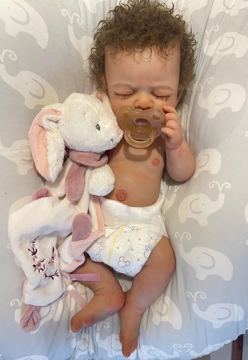May include: A realistic baby doll with curly brown hair, wearing a nappy and holding a dummy. A white and pink plush bunny toy with a scarf is next to the doll. The doll is lying on a grey patterned surface.
