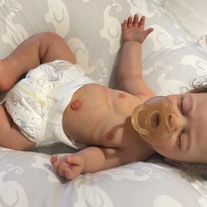 May include: A realistic baby doll with a dummy, wearing a nappy, and lying on a grey patterned blanket. The doll has brown curly hair and is positioned with arms and legs extended.