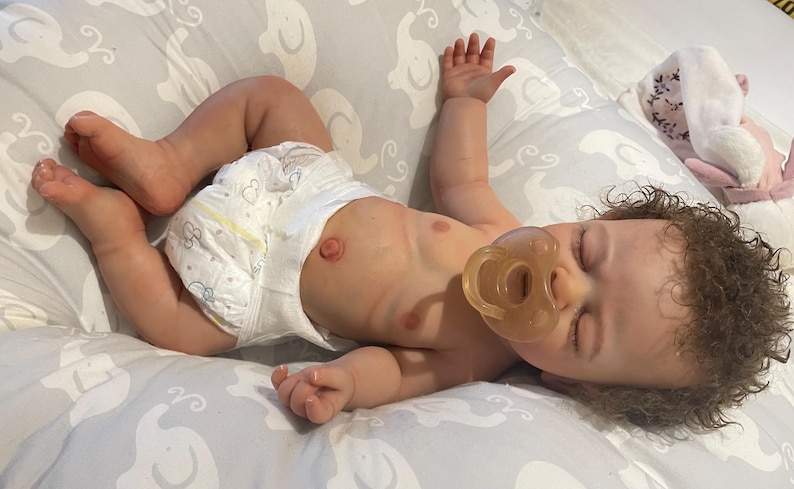 May include: A realistic baby doll with a dummy, wearing a nappy, and lying on a grey patterned blanket. The doll has brown curly hair and is positioned with arms and legs extended.