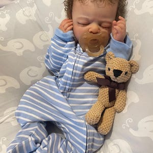 May include: A baby doll wearing a blue and white striped sleepsuit and a dummy. A small, tan teddy bear with a brown ribbon is next to the doll on a grey elephant-patterned blanket.