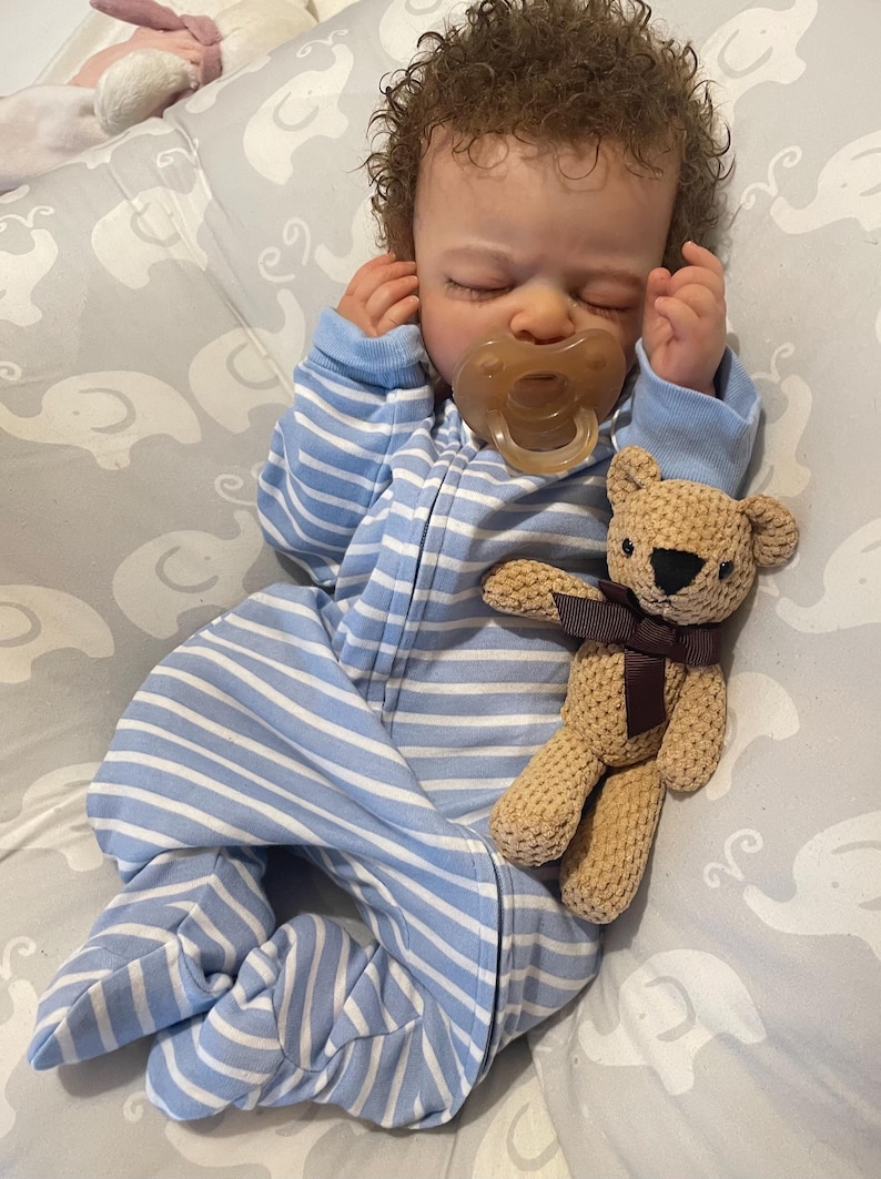 May include: A baby doll wearing a blue and white striped sleepsuit and a dummy. A small, tan teddy bear with a brown ribbon is next to the doll on a grey elephant-patterned blanket.