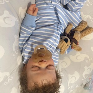 May include: A sleeping baby in a blue and white striped onesie, with a dummy in its mouth. The baby is holding a small, brown teddy bear. The baby has curly brown hair and is lying on a grey patterned blanket.