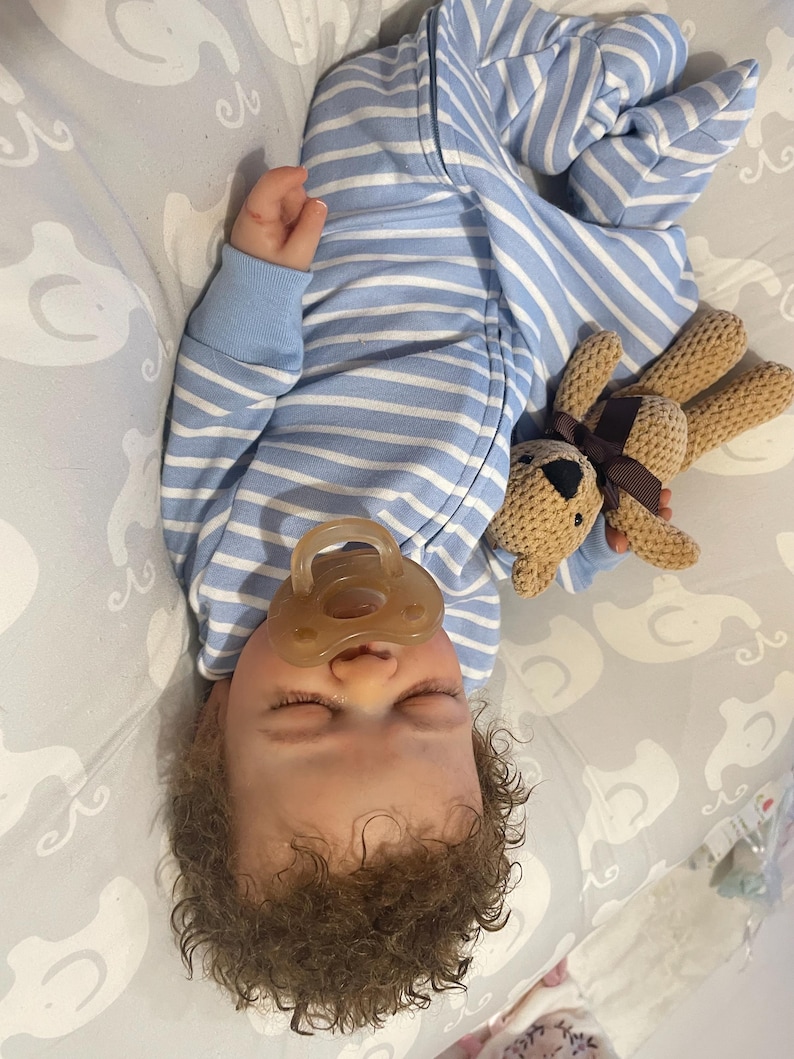 May include: A sleeping baby in a blue and white striped onesie, with a dummy in its mouth. The baby is holding a small, brown teddy bear. The baby has curly brown hair and is lying on a grey patterned blanket.