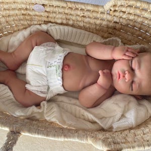 Puede incluir: Una mu&ntilde;eca realista de reci&eacute;n nacido con cabello rizado casta&ntilde;o claro y ojos cerrados, acostada sobre una manta de punto blanca dentro de una cesta de mimbre tejida. La mu&ntilde;eca lleva un pa&ntilde;al desechable blanco y tiene un ombligo detallado.