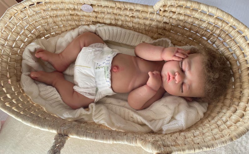 Puede incluir: Una mu&ntilde;eca realista de reci&eacute;n nacido con cabello rizado casta&ntilde;o claro y ojos cerrados, acostada sobre una manta de punto blanca dentro de una cesta de mimbre tejida. La mu&ntilde;eca lleva un pa&ntilde;al desechable blanco y tiene un ombligo detallado.
