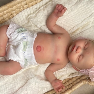 May include: A realistic reborn baby doll with light skin, blonde hair, and closed eyes, wearing a white diaper and a pink lace headband with a bow. The doll is lying on a white knitted blanket inside a woven wicker basket with two handles.