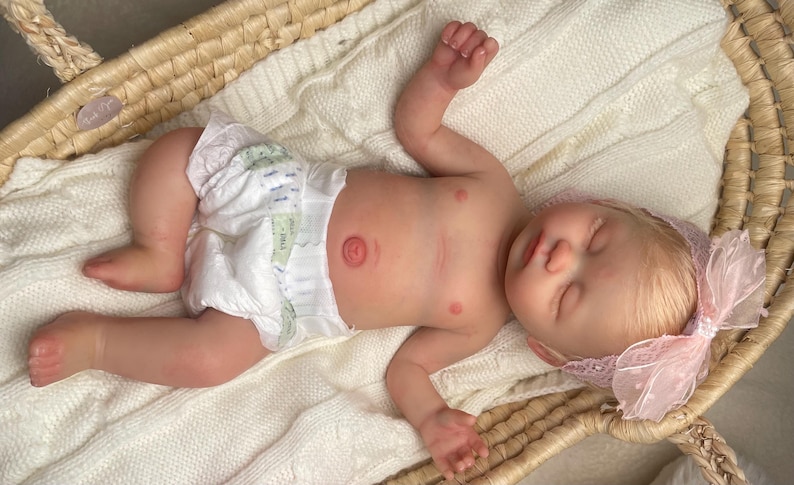 May include: A realistic reborn baby doll with light skin, blonde hair, and closed eyes, wearing a white diaper and a pink lace headband with a bow. The doll is lying on a white knitted blanket inside a woven wicker basket with two handles.