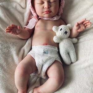 Puede incluir: Una mu&ntilde;eca beb&eacute; realista con un gorro rosa y un pa&ntilde;al, acostada sobre una manta blanca. Un peque&ntilde;o oso de peluche blanco tejido con una cruz negra en la cara descansa junto al beb&eacute;. La mu&ntilde;eca tiene los ojos cerrados y piel clara.