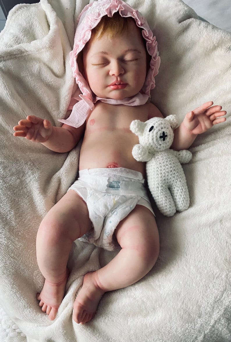 Puede incluir: Una mu&ntilde;eca beb&eacute; realista con un gorro rosa y un pa&ntilde;al, acostada sobre una manta blanca. Un peque&ntilde;o oso de peluche blanco tejido con una cruz negra en la cara descansa junto al beb&eacute;. La mu&ntilde;eca tiene los ojos cerrados y piel clara.