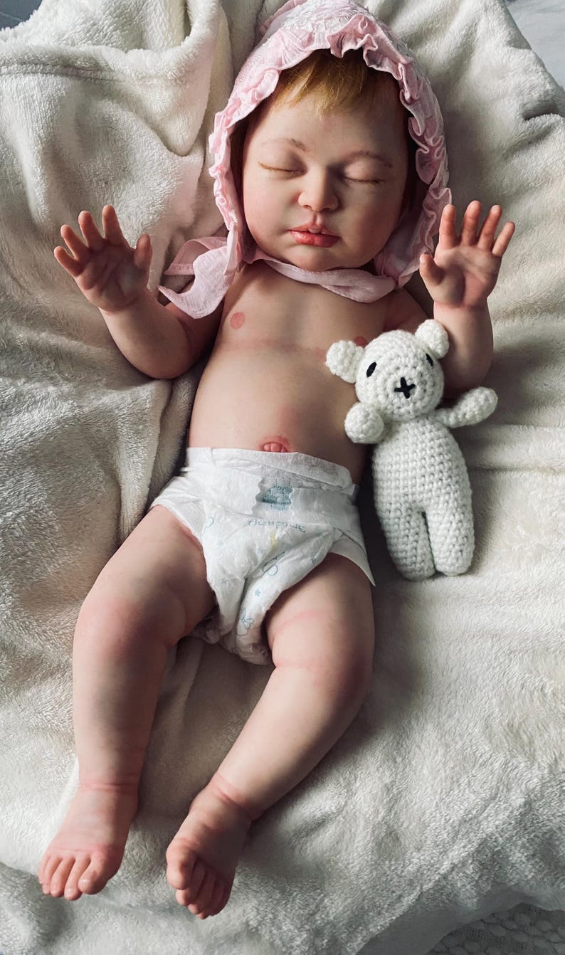 Puede incluir: Una mu&ntilde;eca beb&eacute; realista con un gorro rosa y un pa&ntilde;al, acostada sobre una manta blanca. Un peque&ntilde;o oso de peluche blanco tejido a ganchillo descansa junto a la mu&ntilde;eca. La mu&ntilde;eca tiene los ojos cerrados y piel clara.