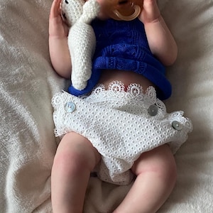 Puede incluir: Mu&ntilde;eca beb&eacute; realista con cabello rojo, vestida con un top azul tejido y un pa&ntilde;al blanco con ribete de encaje. La mu&ntilde;eca sostiene un conejito de peluche blanco y tiene un chupete. Una diadema floral adorna la cabeza de la mu&ntilde;eca.