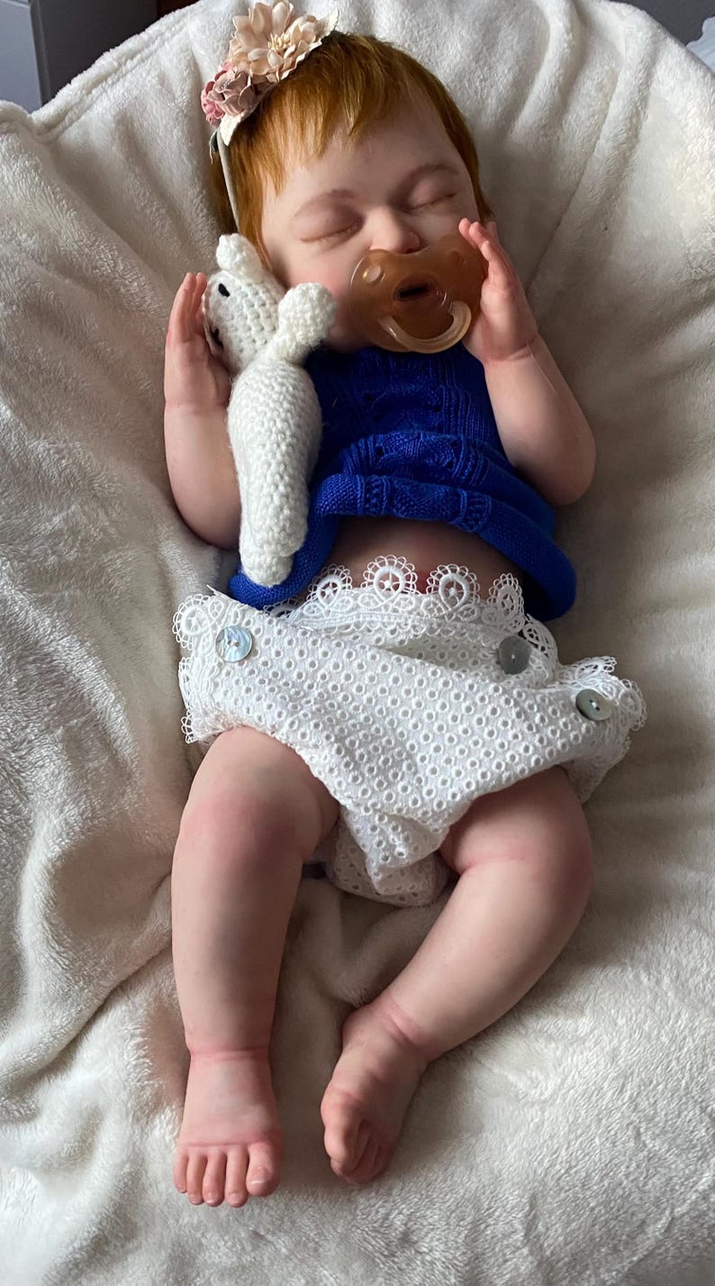 Puede incluir: Mu&ntilde;eca beb&eacute; realista con cabello rojo, vestida con un top azul tejido y un pa&ntilde;al blanco con ribete de encaje. La mu&ntilde;eca sostiene un conejito de peluche blanco y tiene un chupete. Una diadema floral adorna la cabeza de la mu&ntilde;eca.