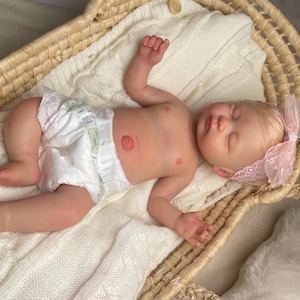 May include: A realistic newborn doll with light skin, blonde hair, and closed eyes, wearing a white diaper and a pink lace headband with a bow. The doll is lying on a white knitted blanket inside a woven wicker basket with two handles.