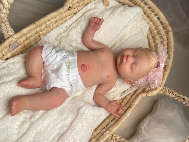 May include: A realistic newborn doll with light skin, blonde hair, and closed eyes, wearing a white diaper and a pink lace headband with a bow. The doll is lying on a white knitted blanket inside a woven wicker basket with two handles.