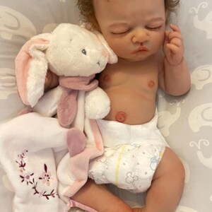 May include: A realistic baby doll with brown curly hair, wearing a nappy, and holding a white plush bunny with pink details. The doll is lying on a grey patterned surface.