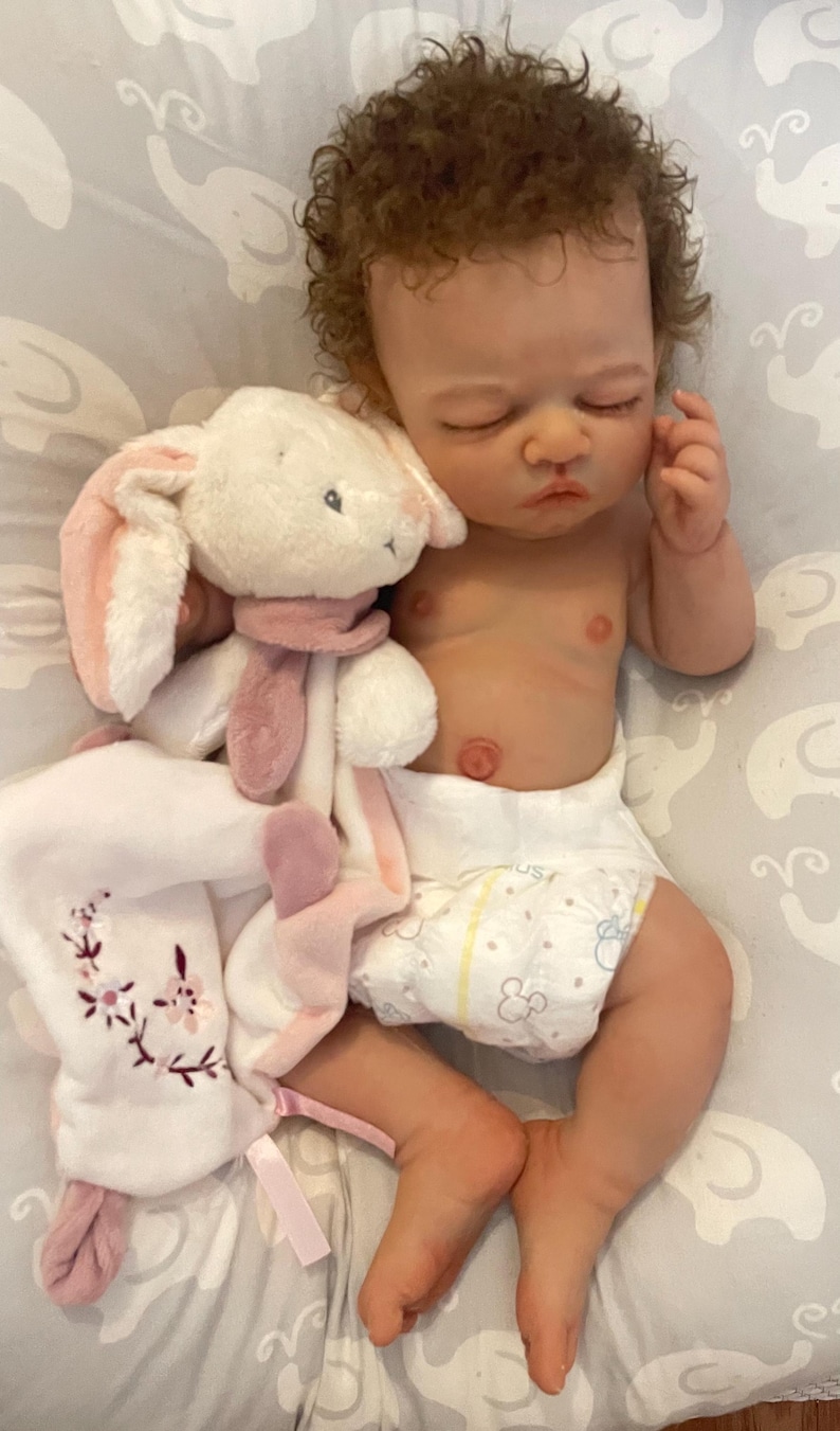 May include: A realistic baby doll with brown curly hair, wearing a nappy, and holding a white plush bunny with pink details. The doll is lying on a grey patterned surface.