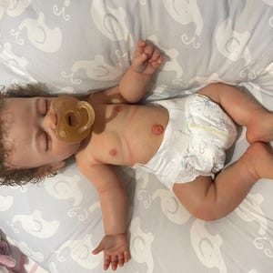 May include: A realistic baby doll with brown curly hair and a dummy in its mouth. The doll is wearing a nappy and is lying on a grey patterned surface. The doll's skin has a realistic tone.