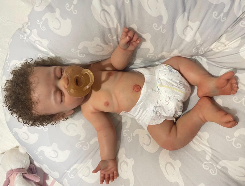 May include: A realistic baby doll with brown curly hair and a dummy in its mouth. The doll is wearing a nappy and is lying on a grey patterned surface. The doll's skin has a realistic tone.