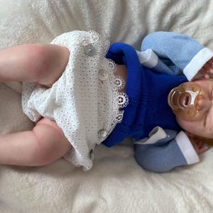 Puede incluir: Una mu&ntilde;eca beb&eacute; realista con un su&eacute;ter azul, pantalones cortos blancos con detalles de botones y zapatos de ganchillo blancos. La mu&ntilde;eca tiene pelo rojo, un chupete y una diadema floral. La mu&ntilde;eca est&aacute; acostada sobre una superficie blanca y suave.