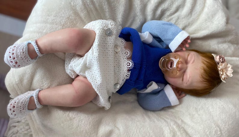 Puede incluir: Una mu&ntilde;eca beb&eacute; realista con un su&eacute;ter azul, pantalones cortos blancos con detalles de botones y zapatos de ganchillo blancos. La mu&ntilde;eca tiene pelo rojo, un chupete y una diadema floral. La mu&ntilde;eca est&aacute; acostada sobre una superficie blanca y suave.