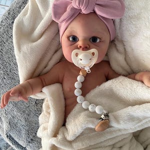 May include: A realistic baby doll with blue eyes, wearing a pink knotted headband and a white pacifier with a wooden beaded chain.