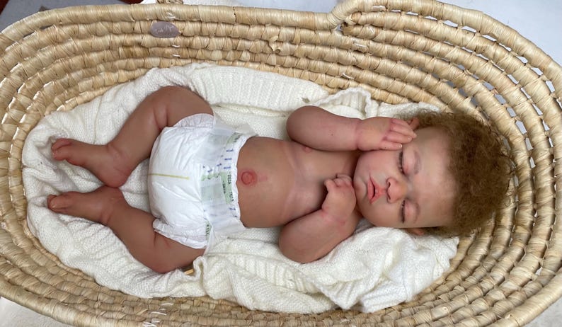 Puede incluir: Una mu&ntilde;eca reci&eacute;n nacida realista con piel clara, cabello casta&ntilde;o rizado y ojos cerrados, que lleva un pa&ntilde;al desechable blanco. La mu&ntilde;eca est&aacute; acostada sobre una manta de punto blanca dentro de una cesta de mimbre ovalada de color marr&oacute;n.