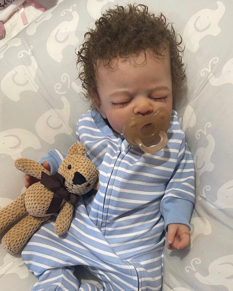 May include: A baby doll with brown curly hair, wearing a blue and white striped onesie and a dummy. The doll is holding a small, tan teddy bear with a brown bow.