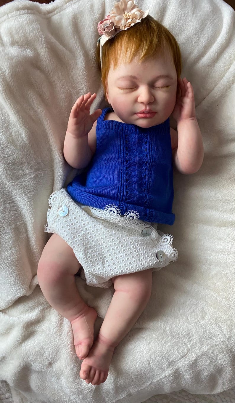 Puede incluir: Una mu&ntilde;eca beb&eacute; realista con cabello rojo y ojos cerrados, vestida con un top de punto azul y pantalones cortos de encaje blanco. La mu&ntilde;eca tiene una diadema floral y est&aacute; acostada sobre una suave manta blanca.