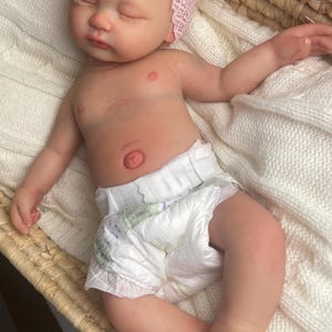 May include: A realistic newborn baby doll with light skin and blonde hair, wearing a white diaper and a pink lace headband with a bow. The doll is lying down with eyes closed, featuring detailed skin texture, a navel, and small fingers and toes.