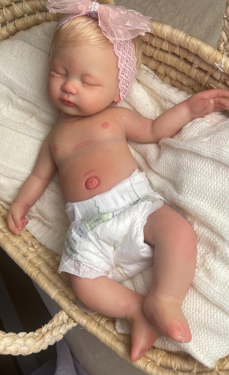 May include: A realistic newborn baby doll with light skin and blonde hair, wearing a white diaper and a pink lace headband with a bow. The doll is lying down with eyes closed, featuring detailed skin texture, a navel, and small fingers and toes.