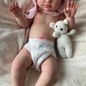 Puede incluir: Una mu&ntilde;eca beb&eacute; realista con piel clara, que lleva un gorro rosa y un pa&ntilde;al. Un peque&ntilde;o oso de peluche blanco de ganchillo est&aacute; al lado de la mu&ntilde;eca. La mu&ntilde;eca est&aacute; tumbada sobre una manta blanca texturizada.