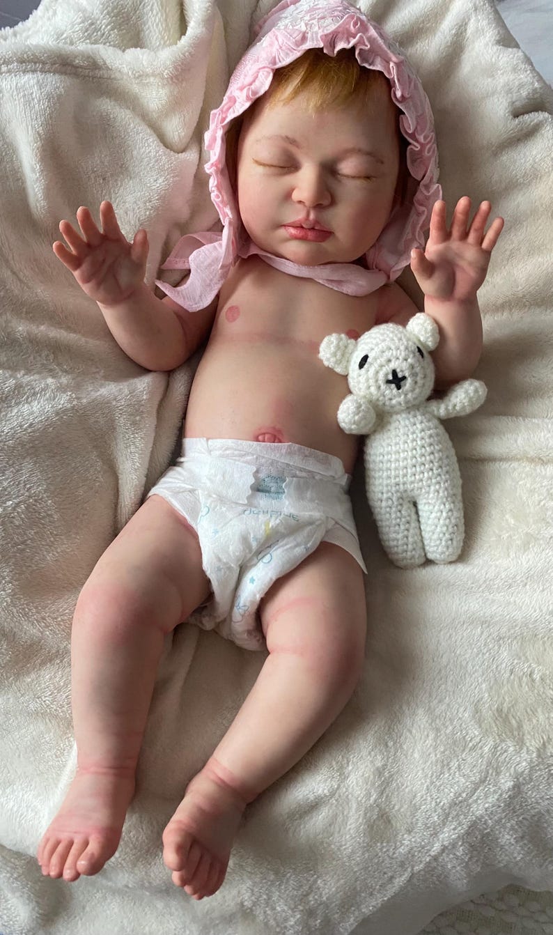 Puede incluir: Una mu&ntilde;eca beb&eacute; realista con piel clara, que lleva un gorro rosa y un pa&ntilde;al. Un peque&ntilde;o oso de peluche blanco de ganchillo est&aacute; al lado de la mu&ntilde;eca. La mu&ntilde;eca est&aacute; tumbada sobre una manta blanca texturizada.