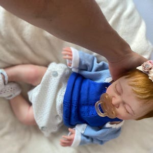 Puede incluir: Una mu&ntilde;eca beb&eacute; realista con un su&eacute;ter azul, pantalones cortos blancos y botines de ganchillo. La mu&ntilde;eca tiene el pelo rojo y una diadema floral. Un chupete est&aacute; en su boca.