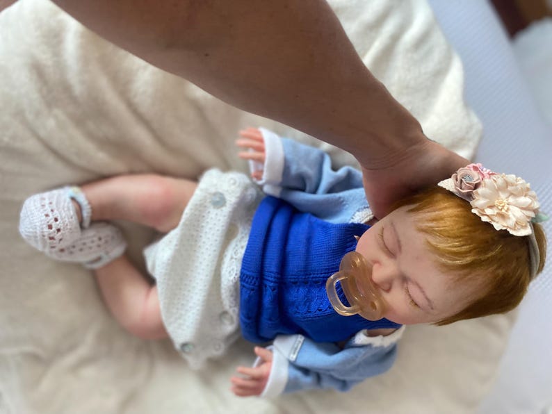 Puede incluir: Una mu&ntilde;eca beb&eacute; realista con un su&eacute;ter azul, pantalones cortos blancos y botines de ganchillo. La mu&ntilde;eca tiene el pelo rojo y una diadema floral. Un chupete est&aacute; en su boca.