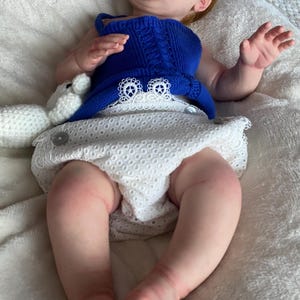 Puede incluir: Una mu&ntilde;eca beb&eacute; realista con piel clara, cabello rojo y ojos cerrados. La mu&ntilde;eca lleva un top azul tejido y una cubierta de pa&ntilde;al blanca con ojales. Un peluche blanco est&aacute; al lado de la mu&ntilde;eca.