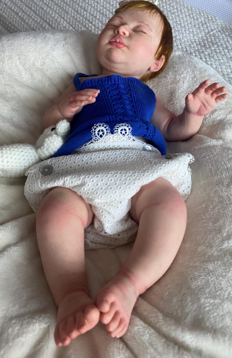 Puede incluir: Una mu&ntilde;eca beb&eacute; realista con piel clara, cabello rojo y ojos cerrados. La mu&ntilde;eca lleva un top azul tejido y una cubierta de pa&ntilde;al blanca con ojales. Un peluche blanco est&aacute; al lado de la mu&ntilde;eca.