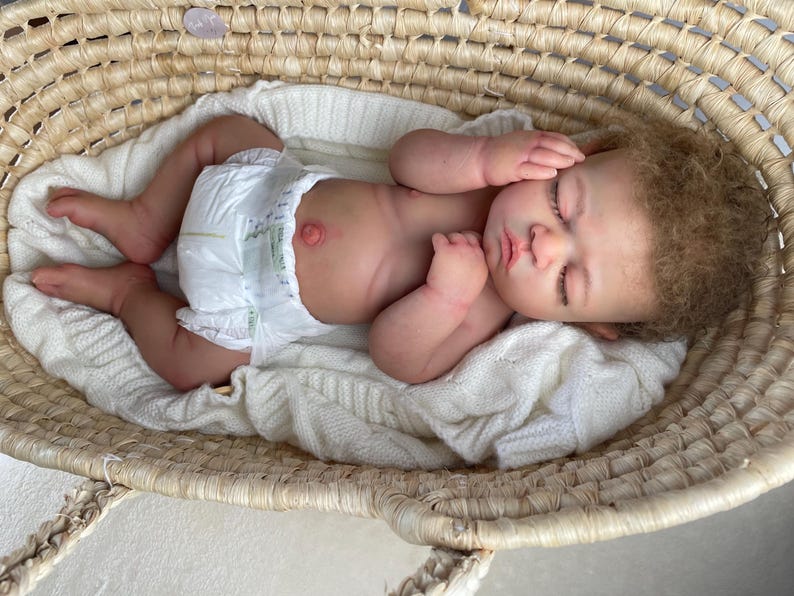 Puede incluir: Una mu&ntilde;eca realista de reci&eacute;n nacido con piel clara, ojos cerrados y cabello casta&ntilde;o rizado, que lleva un pa&ntilde;al desechable blanco. La mu&ntilde;eca est&aacute; acostada sobre una manta de punto blanca dentro de una cesta de mimbre marr&oacute;n claro con dos asas.
