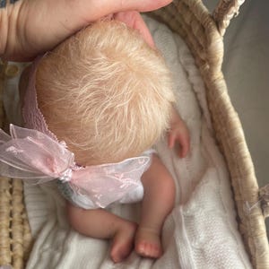 May include: A realistic baby doll with fine blonde hair, wearing a pink lace headband featuring a sheer organza bow. The doll is positioned inside a woven wicker basket lined with a white knitted blanket, showcasing detailed skin tones and small limbs.