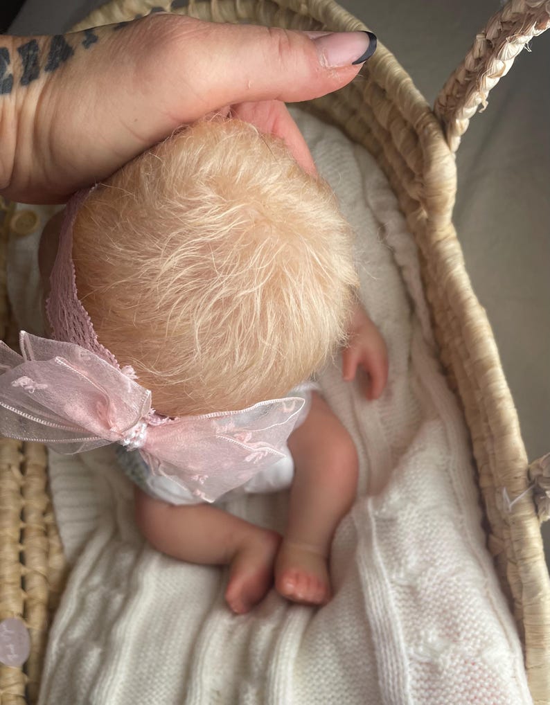 May include: A realistic baby doll with fine blonde hair, wearing a pink lace headband featuring a sheer organza bow. The doll is positioned inside a woven wicker basket lined with a white knitted blanket, showcasing detailed skin tones and small limbs.