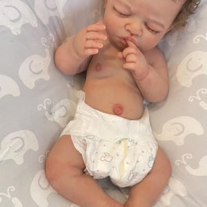 May include: A lifelike baby doll with closed eyes and a nappy. The doll has detailed skin tones, brown curly hair, and is lying on a grey patterned blanket. The doll's hands are near its face.