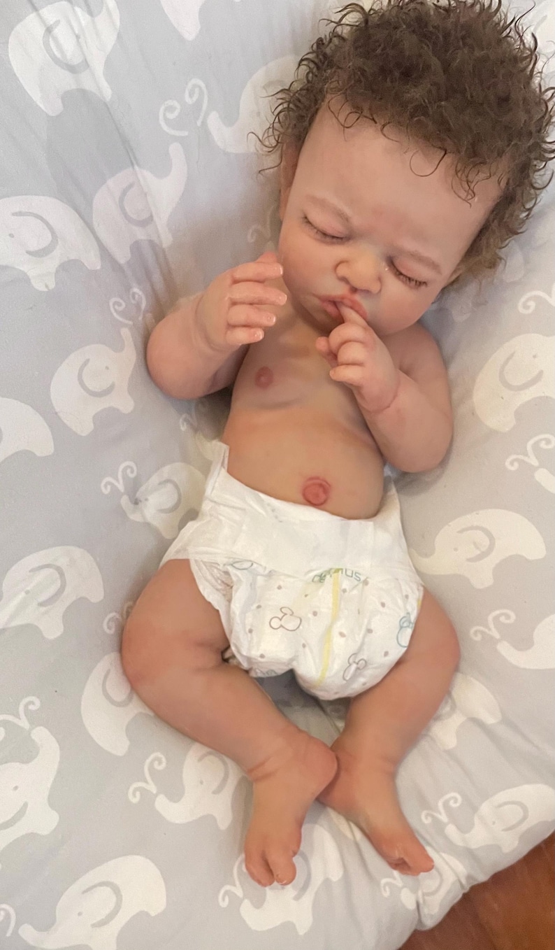 May include: A lifelike baby doll with closed eyes and a nappy. The doll has detailed skin tones, brown curly hair, and is lying on a grey patterned blanket. The doll's hands are near its face.