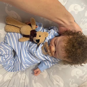 May include: A baby in blue and white striped pyjamas, with a dummy in its mouth, sleeps peacefully. A small, tan teddy bear with a brown ribbon rests on the baby. The baby has curly brown hair.
