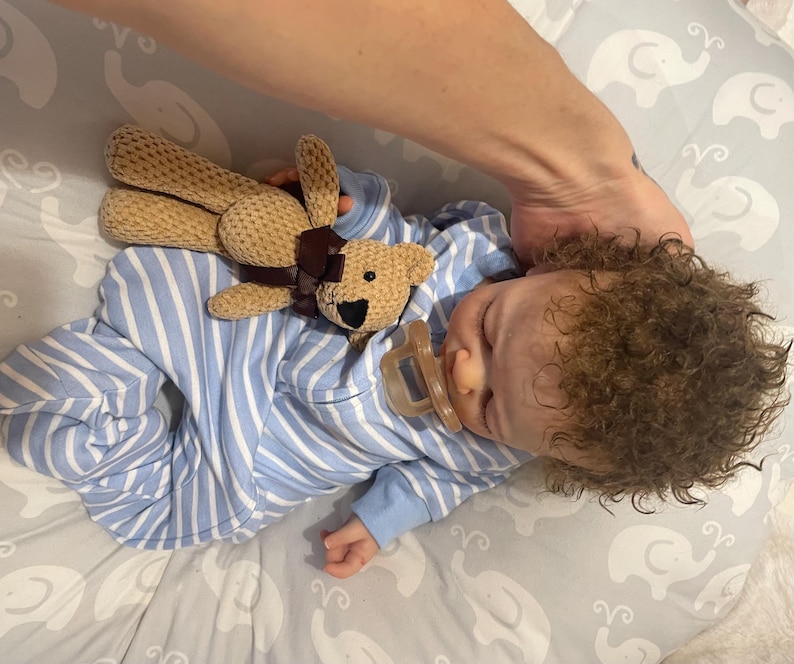 May include: A baby in blue and white striped pyjamas, with a dummy in its mouth, sleeps peacefully. A small, tan teddy bear with a brown ribbon rests on the baby. The baby has curly brown hair.