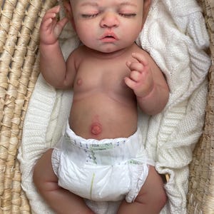 Puede incluir: Una mu&ntilde;eca reborn realista con cabello rizado casta&ntilde;o claro y ojos cerrados, acostada boca arriba. La mu&ntilde;eca tiene un tono de piel detallado, un ombligo prominente y lleva un pa&ntilde;al desechable blanco. Est&aacute; colocada dentro de una cesta.