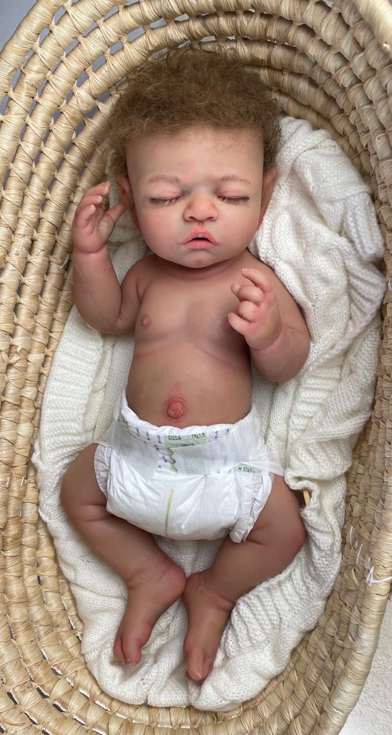Puede incluir: Una mu&ntilde;eca reborn realista con cabello rizado casta&ntilde;o claro y ojos cerrados, acostada boca arriba. La mu&ntilde;eca tiene un tono de piel detallado, un ombligo prominente y lleva un pa&ntilde;al desechable blanco. Est&aacute; colocada dentro de una cesta.